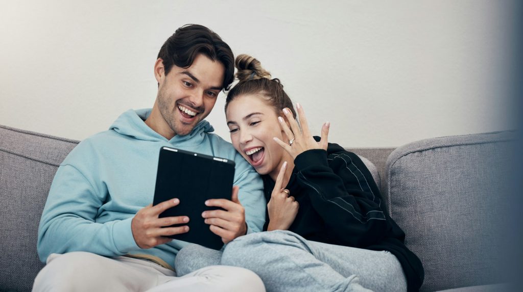 Man, woman and video call for engagement ring with happiness on sofa in living room of home. People